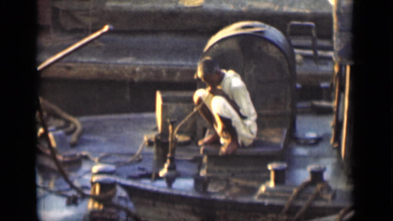 Man in white clothing crouches on a boat deck near ropes and a large cylindrical tank.