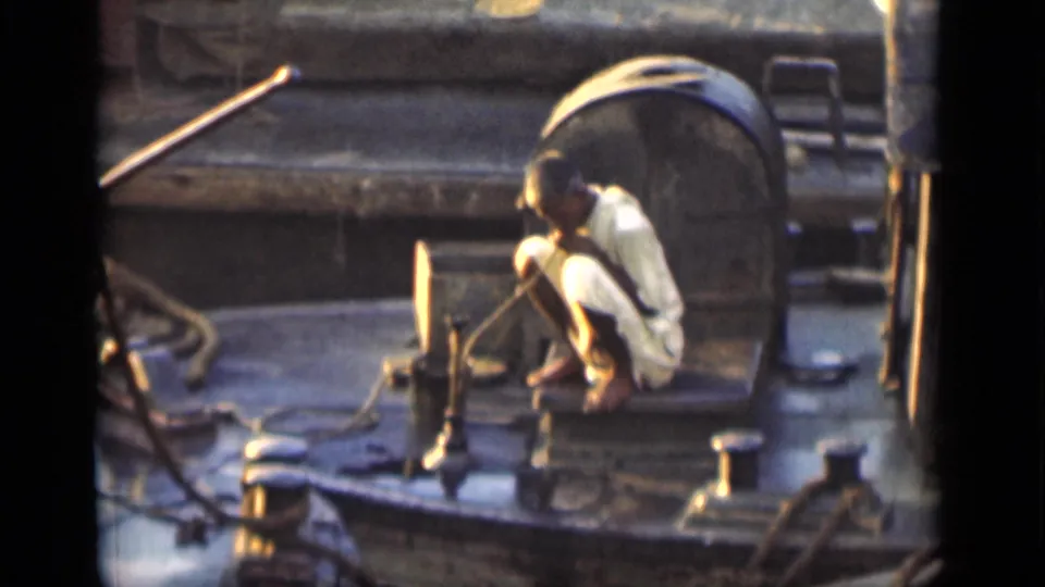 Man in white clothing crouches on a Bombay boat deck near hoses, ropes, fittings, and a large cylindrical tank.