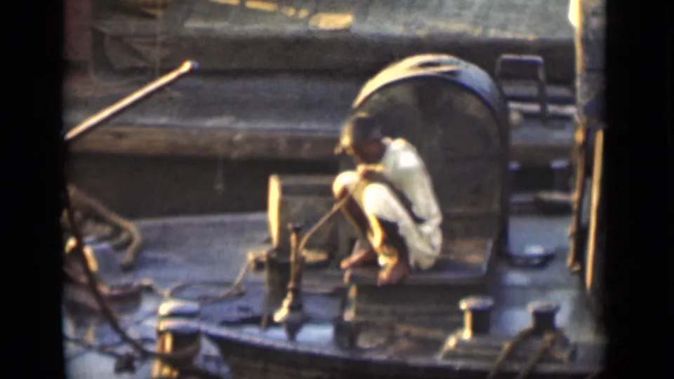 Crouched worker on a Bombay boat deck near a large cylindrical tank, ropes, posts, and sunlit maritime hardware.