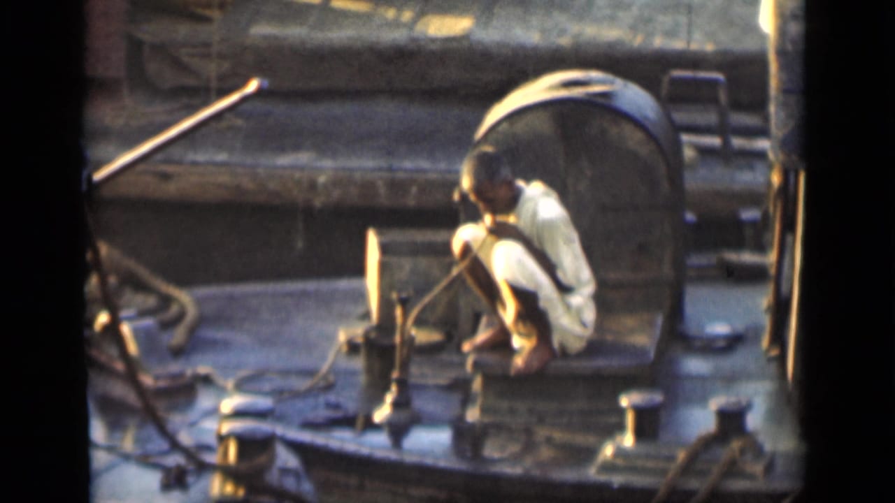 Man crouched on a boat deck beside ropes and metal fittings in warm sunlight.
