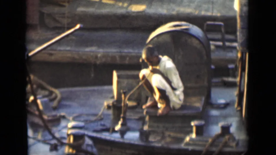 In Bombay home-movie footage from 1946, a man crouches on a boat deck near ropes, posts, and metal fixtures.