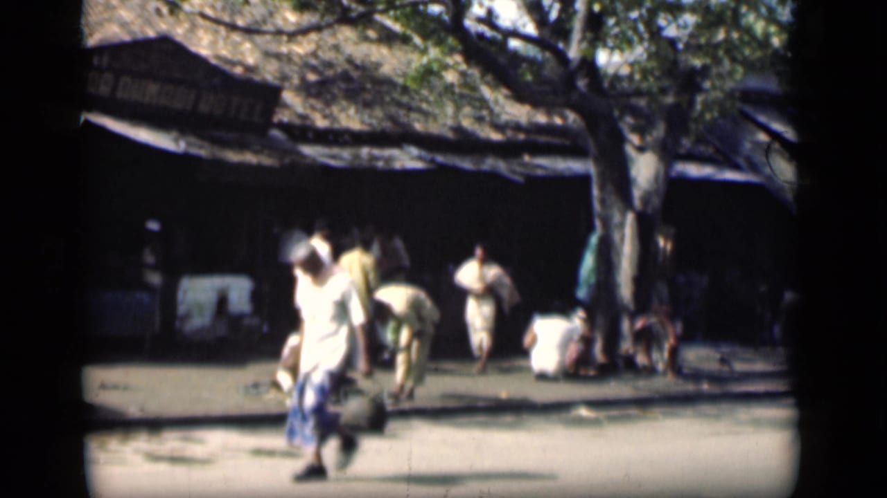 Pedestrians pass a shaded street frontage with a painted hotel sign and a large tree.