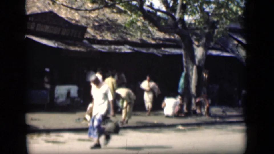 Bombay street scene from 1946 with pedestrians moving past a shaded market or hotel frontage beneath a large tree.
