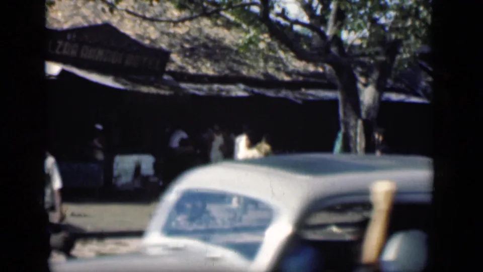A vintage car crosses the foreground of a 1946 Bombay street view while people stand near a shaded frontage.