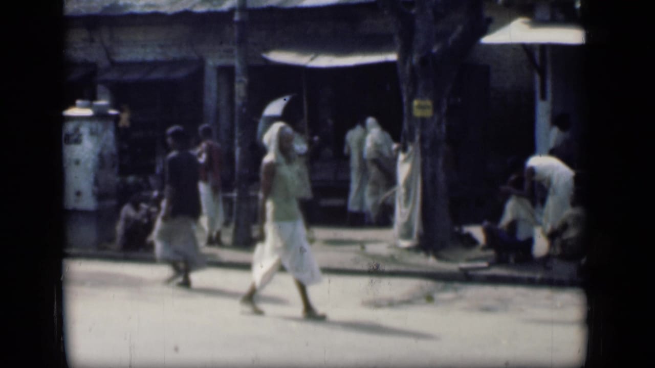 1946 Bombay street view with a woman in a white sari crossing in front of shops, seated figures, and shaded roadside activity.