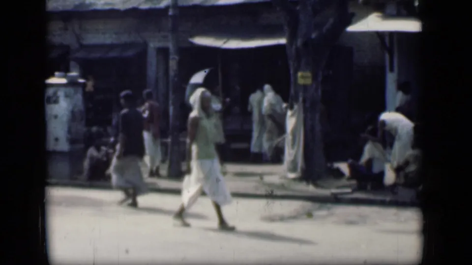 1946 Bombay street view with a woman in a white sari crossing in front of shops, seated figures, and shaded roadside activity.