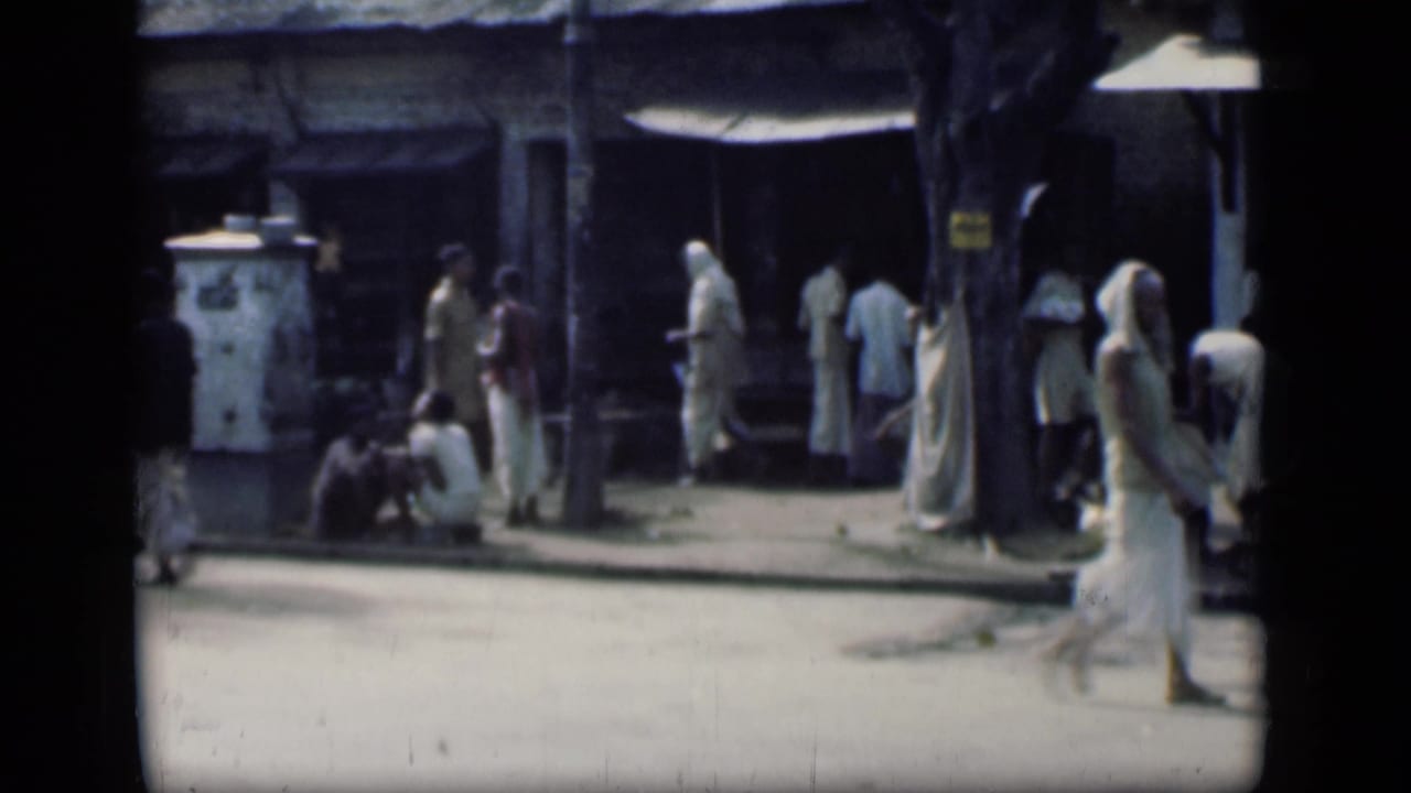People stand and sit near shopfronts as a sari-clad pedestrian exits frame.