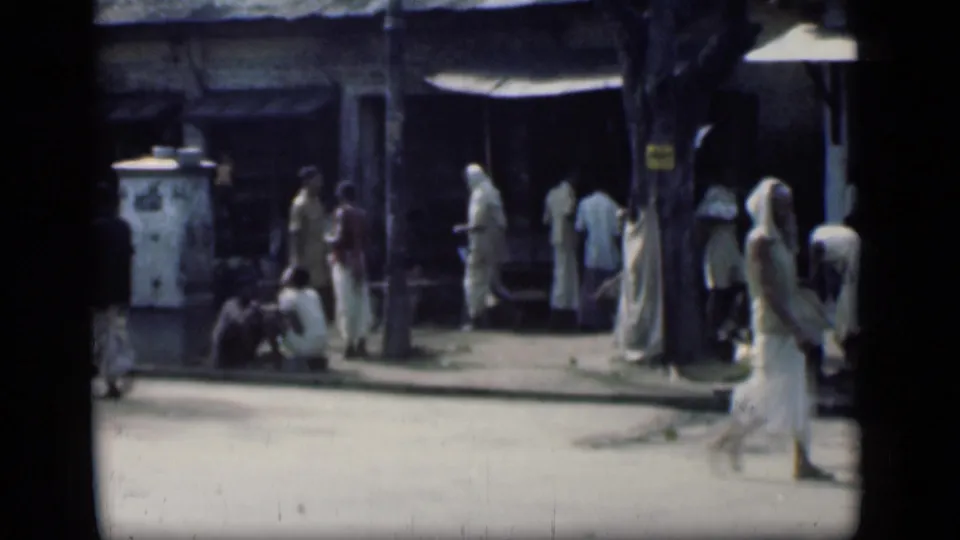 1946 Bombay street scene with small groups near shaded shops, seated people by the curb, and pedestrians moving through bright light.