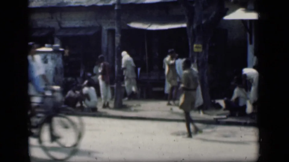 A bicycle moves through the foreground of a 1946 Bombay street view as pedestrians and seated figures remain near shopfronts.