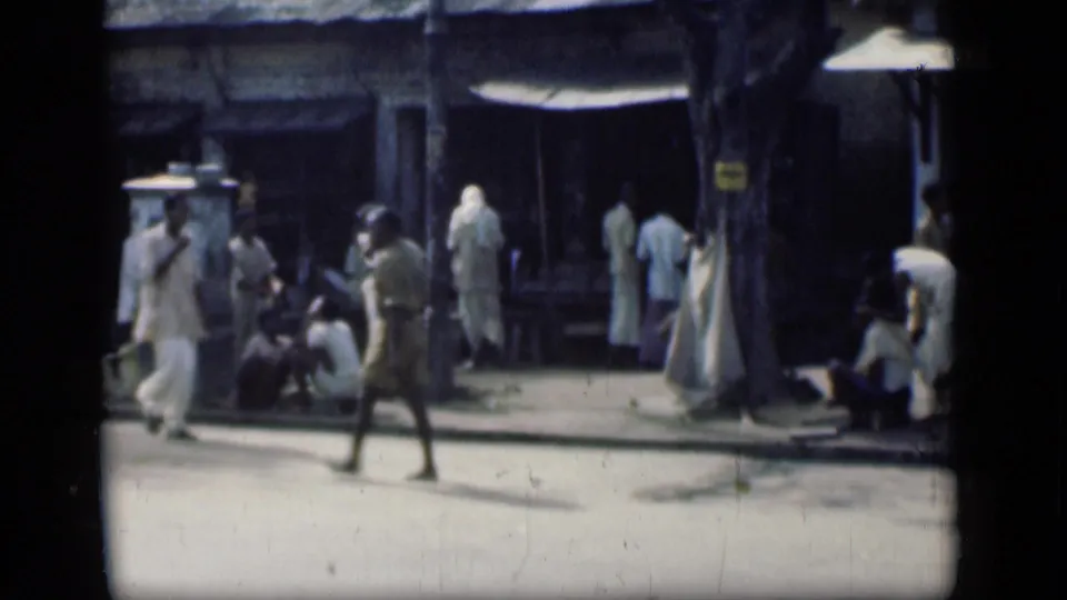 1946 Bombay street frame with a man crossing the sunlit roadway and several people standing or sitting near shaded shopfronts.