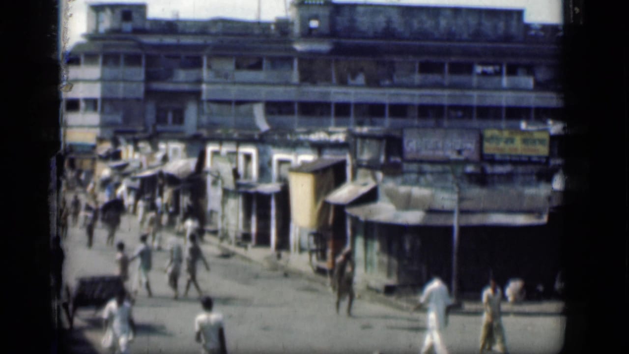 People move along a street with shop awnings and a large building behind them.