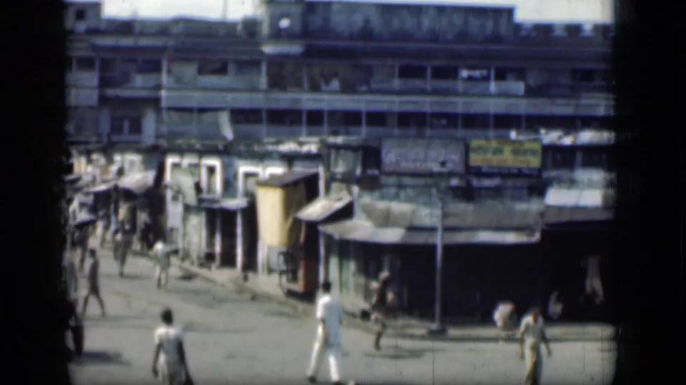 Home-movie frame of 1946 Bombay showing a street corner, pedestrians, a handcart, small shops and a worn multi-story facade.