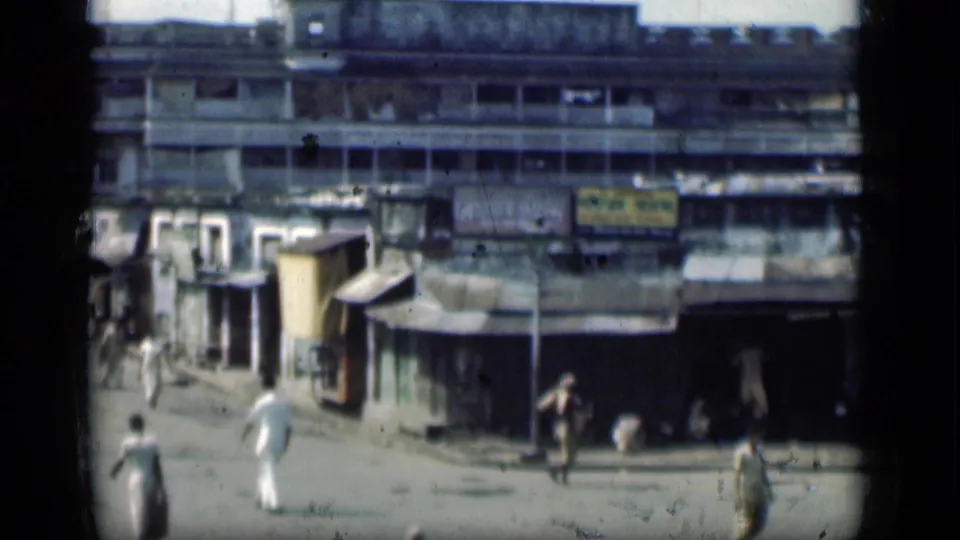 1946 Bombay street view with pedestrians, shaded shopfronts, visible signs and a large weathered building across the background.