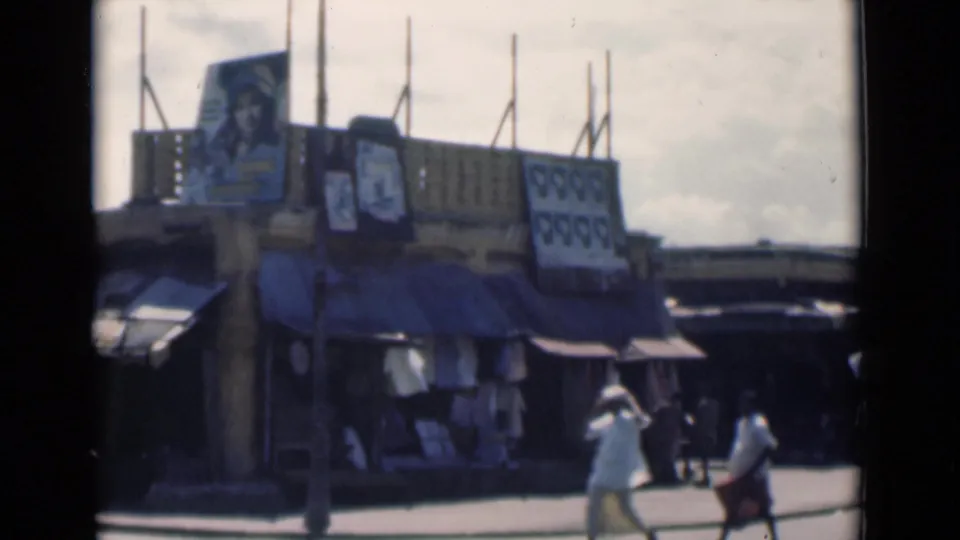 1946 Calcutta street scene with pedestrians in period clothing moving beside awnings, storefronts, and large posted images.