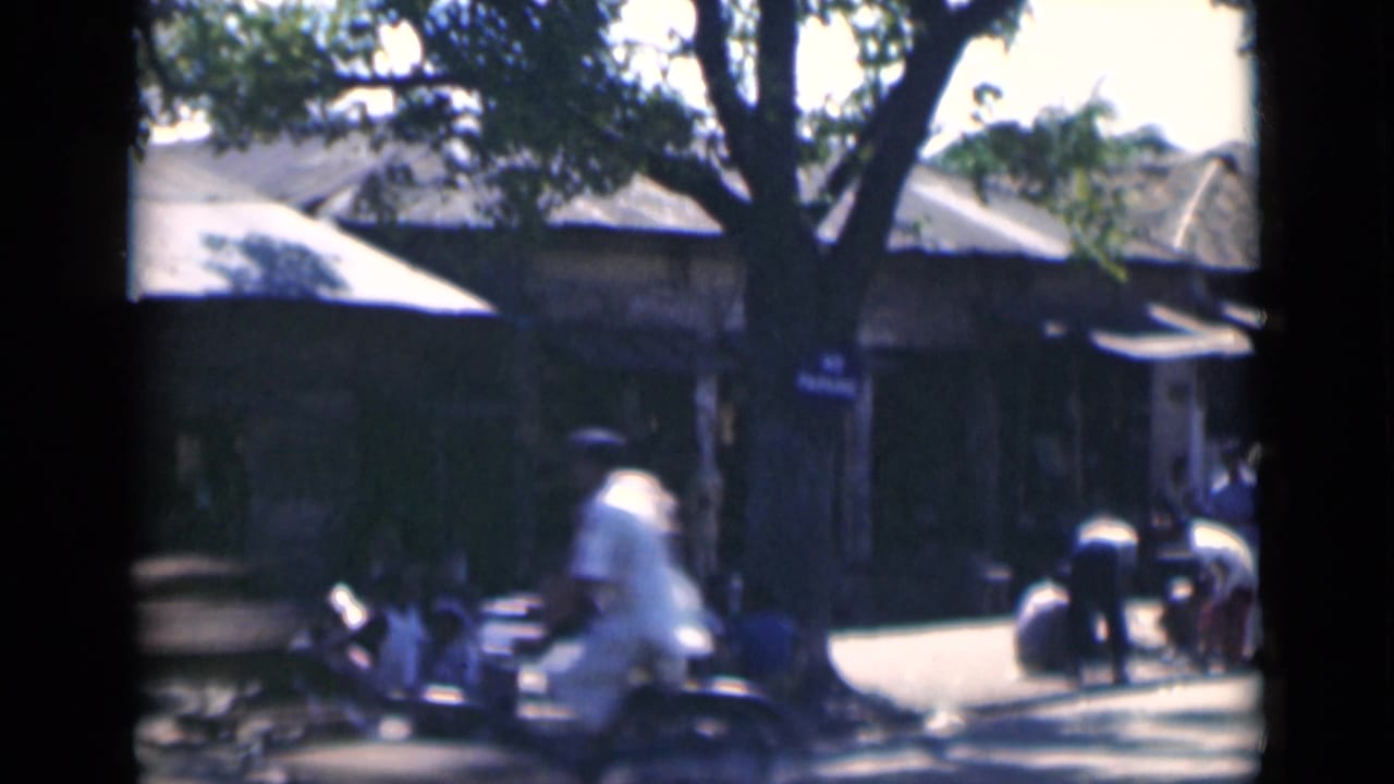 Street corner with pedestrians, shade trees, low buildings, and a person riding or sitting near a bicycle.