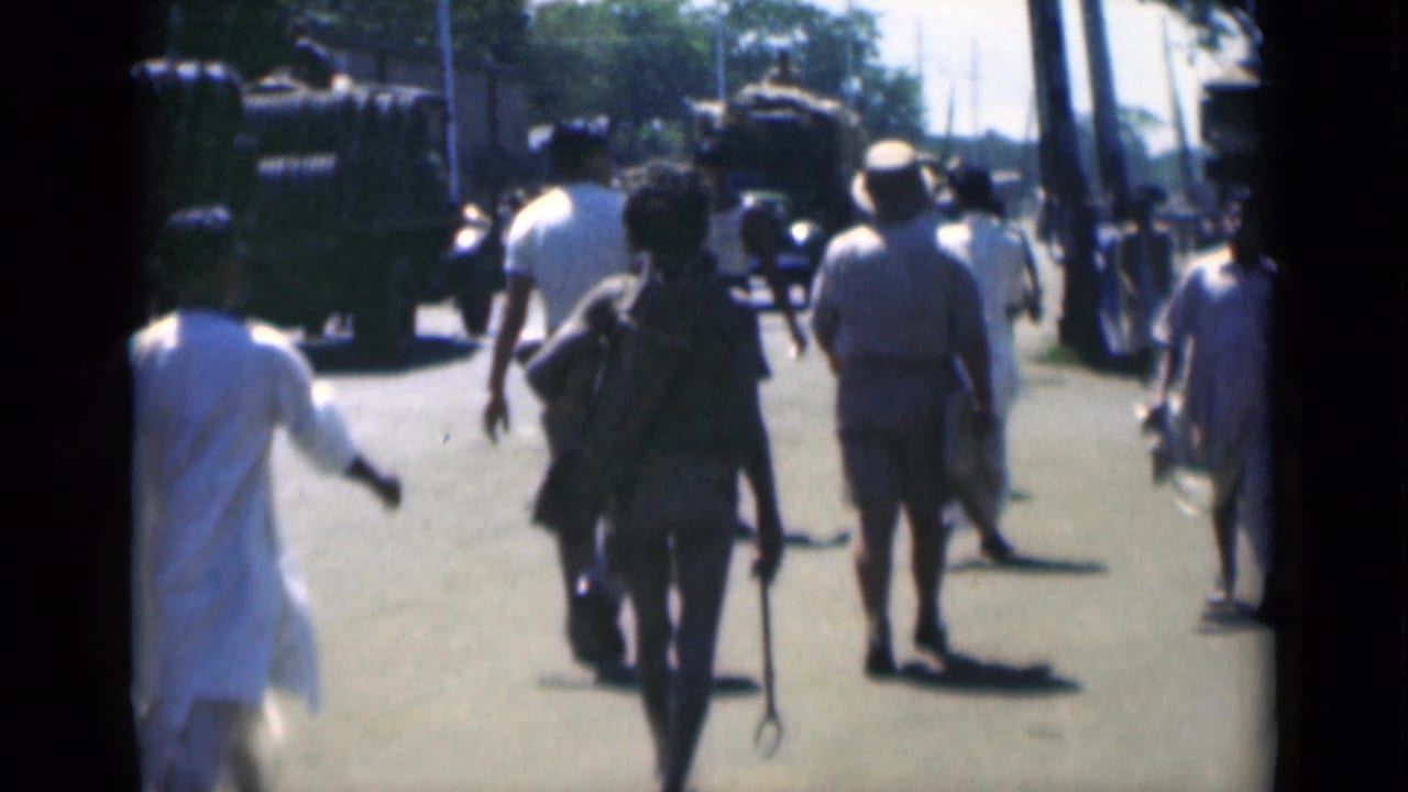People walking along a bright street with large military-style vehicles ahead and trees along the road.