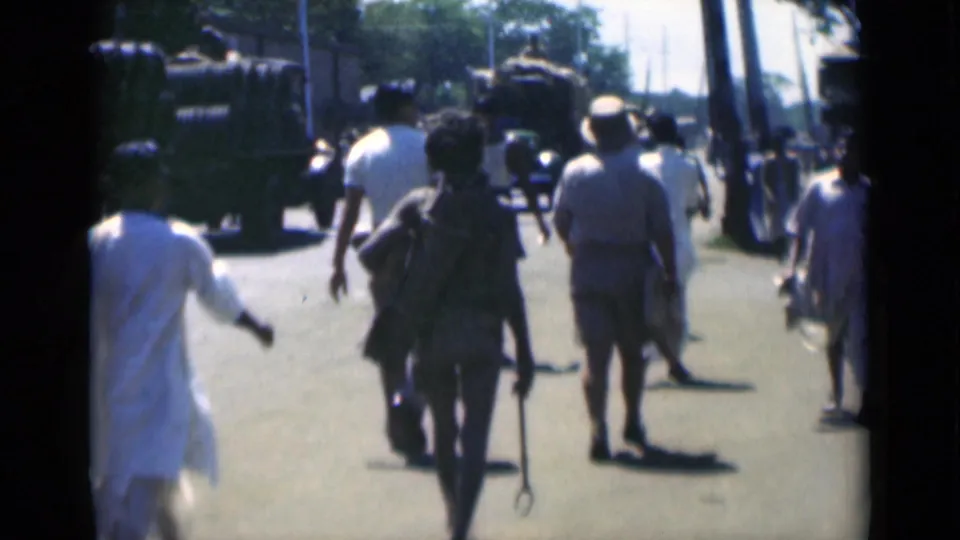 1946 Calcutta street scene with several pedestrians walking away from the camera toward large military-style vehicles on a sunlit road.