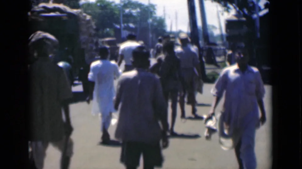 1946 Calcutta street footage showing pedestrians walking away from the camera near palm trunks, roadside vehicles, and bright shadows.