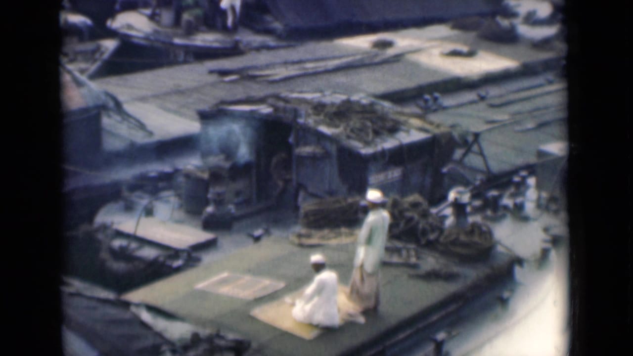 Two men in white clothing on prayer rugs on a boat deck with ropes and cargo nearby.