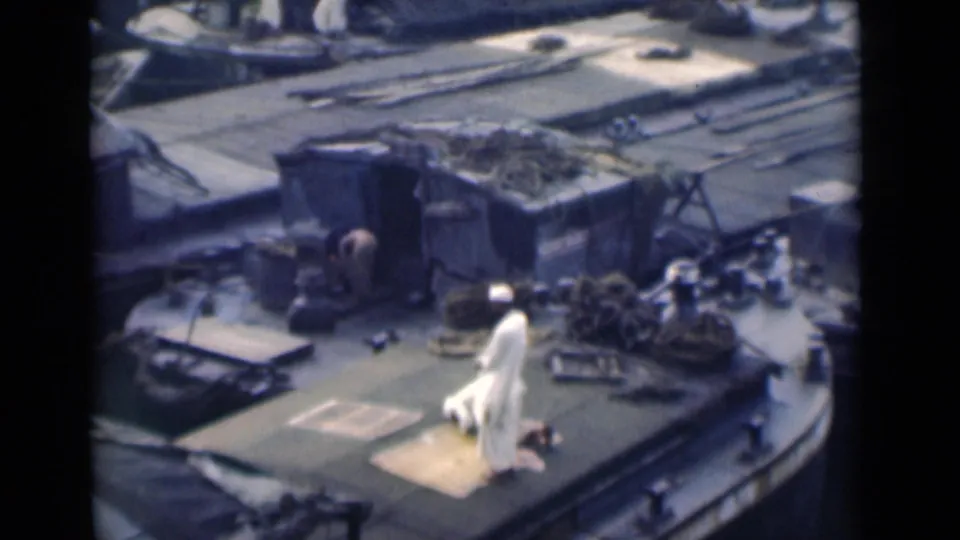 A man in white kneels on a prayer rug on a working boat deck in the 1946 Calcutta footage, with rope and cargo nearby.