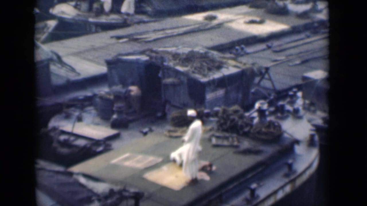 Man in white clothing praying on a rug on a ship deck near ropes and metal fixtures.