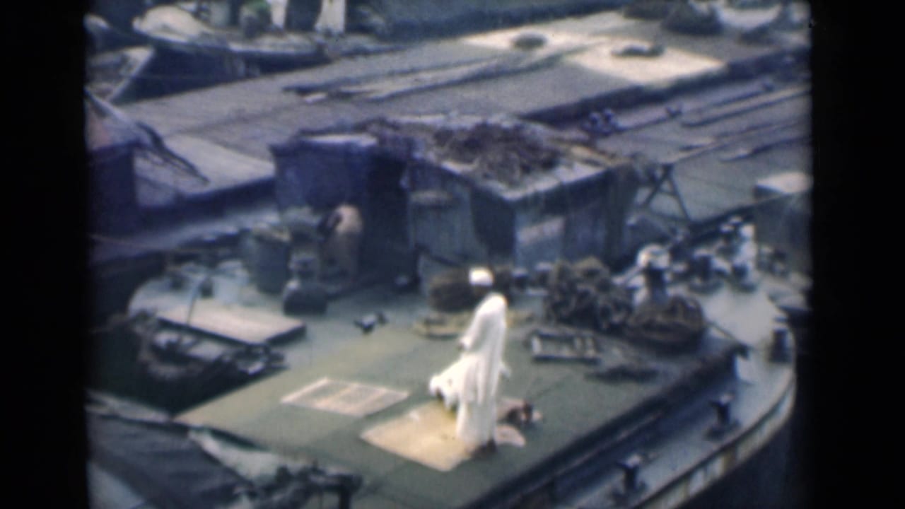 A Muslim man in white clothing continues prayer on a rug on a ship deck, with ropes and cargo-like fixtures nearby.