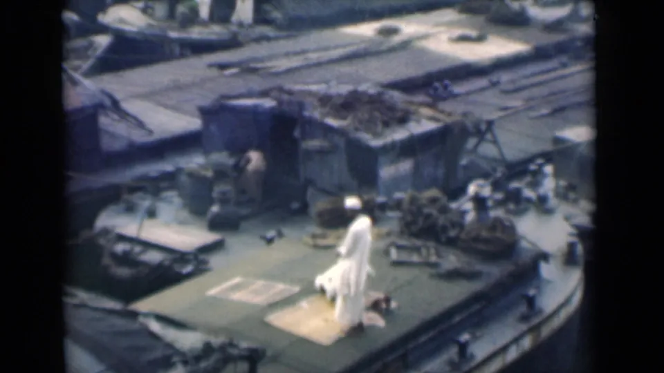 A Muslim man in white clothing continues prayer on a rug on a ship deck, with ropes and cargo-like fixtures nearby.
