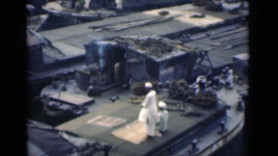 A Muslim man bends forward in prayer on a rug aboard a ship, with ropes, railings, and deck fixtures visible around him.