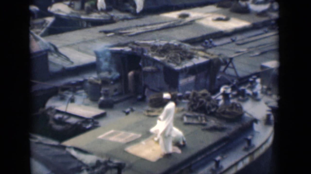 In 1946 Calcutta home-movie footage, two men in white clothing appear on a boat deck as one kneels on a mat near ropes and deck fittings.