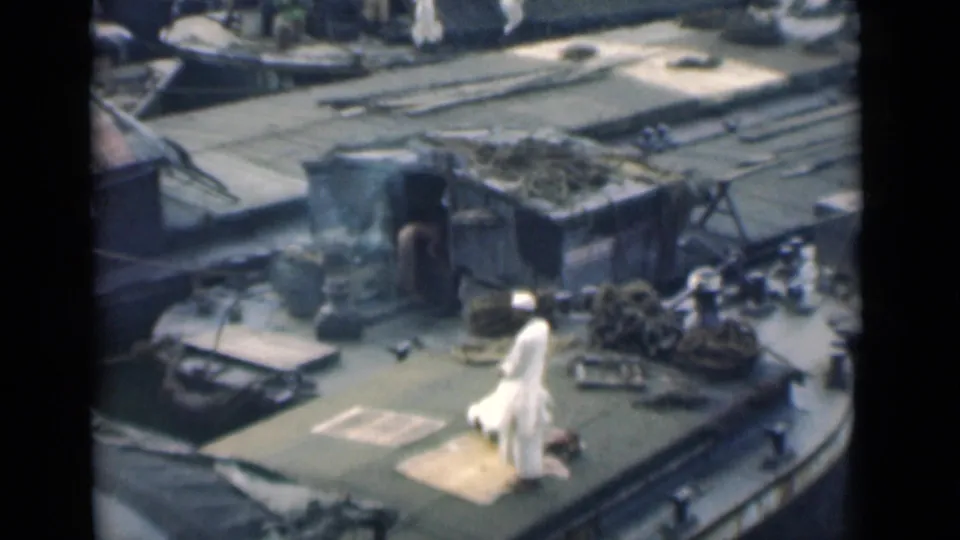 A high-angle 8mm frame from Calcutta shows two men on the deck, one standing and one kneeling near a mat amid rope coils and vessel hardware.