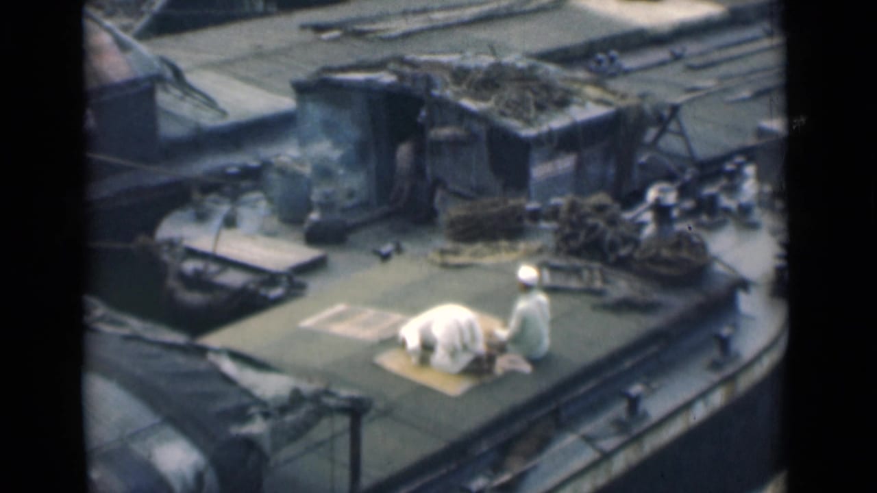 Two men in white clothing on a boat deck, one kneeling on a mat while the other stands nearby.