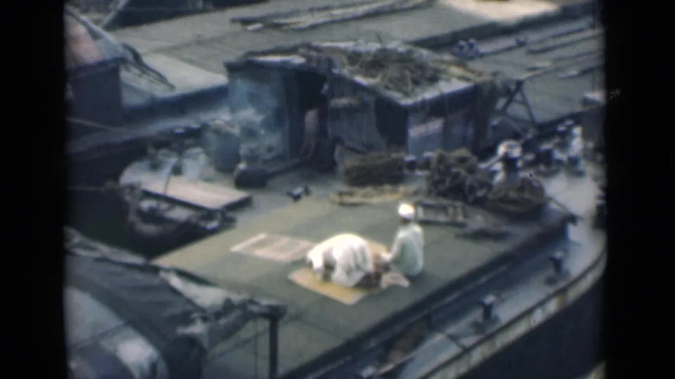 In 1946 Calcutta footage, two Muslim men appear on a cluttered boat deck as one kneels on a prayer mat and the other stands beside him.