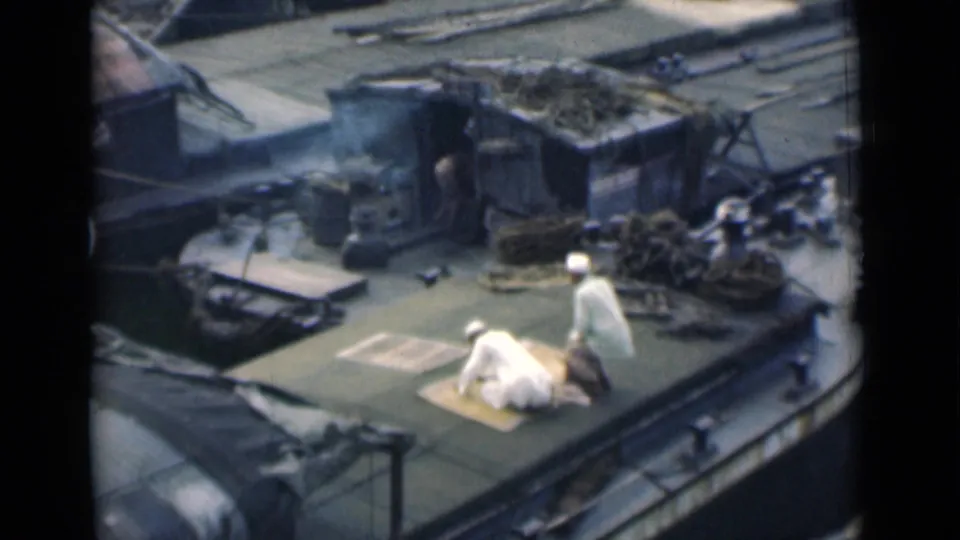 In the 1946 Calcutta home-movie frame, a worshipper is low on the mat while a second man stands beside him on the crowded boat deck.