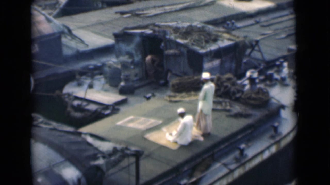 Two men in white clothing on a rooftop terrace, one kneeling on a mat while the other stands nearby.