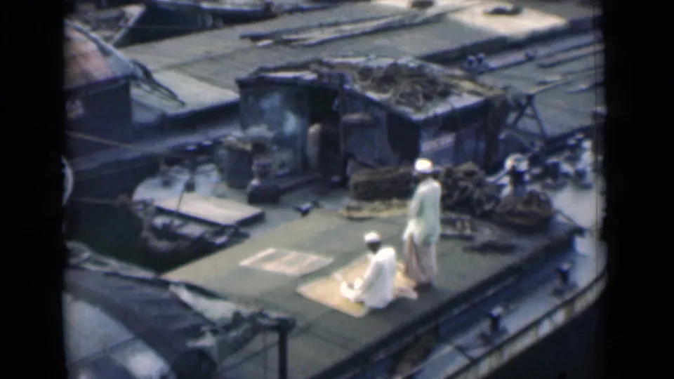 1946 Calcutta 8mm frame showing two men in white garments beside a prayer mat on a cluttered rooftop terrace.