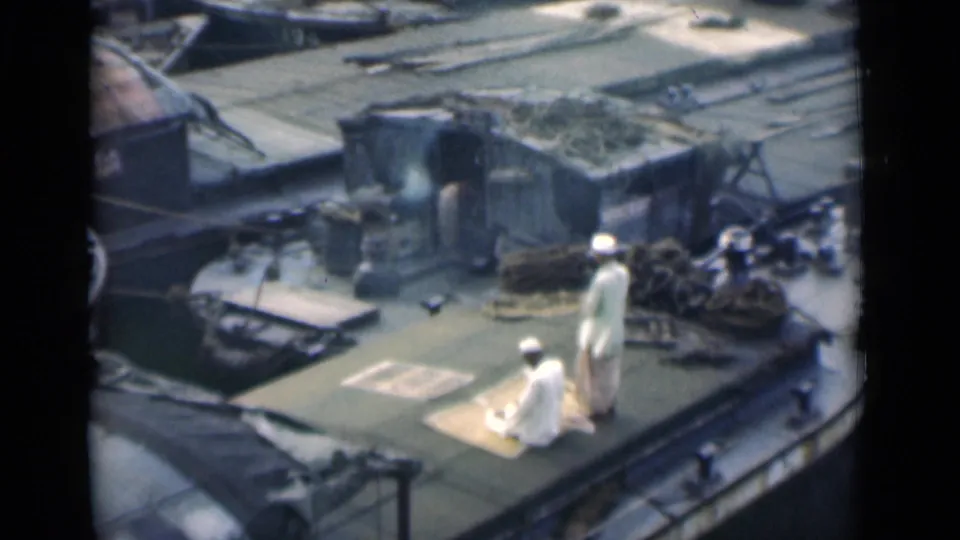 1946 Calcutta home-movie still of two Muslim men on a rooftop prayer mat area, filmed from an elevated distance.