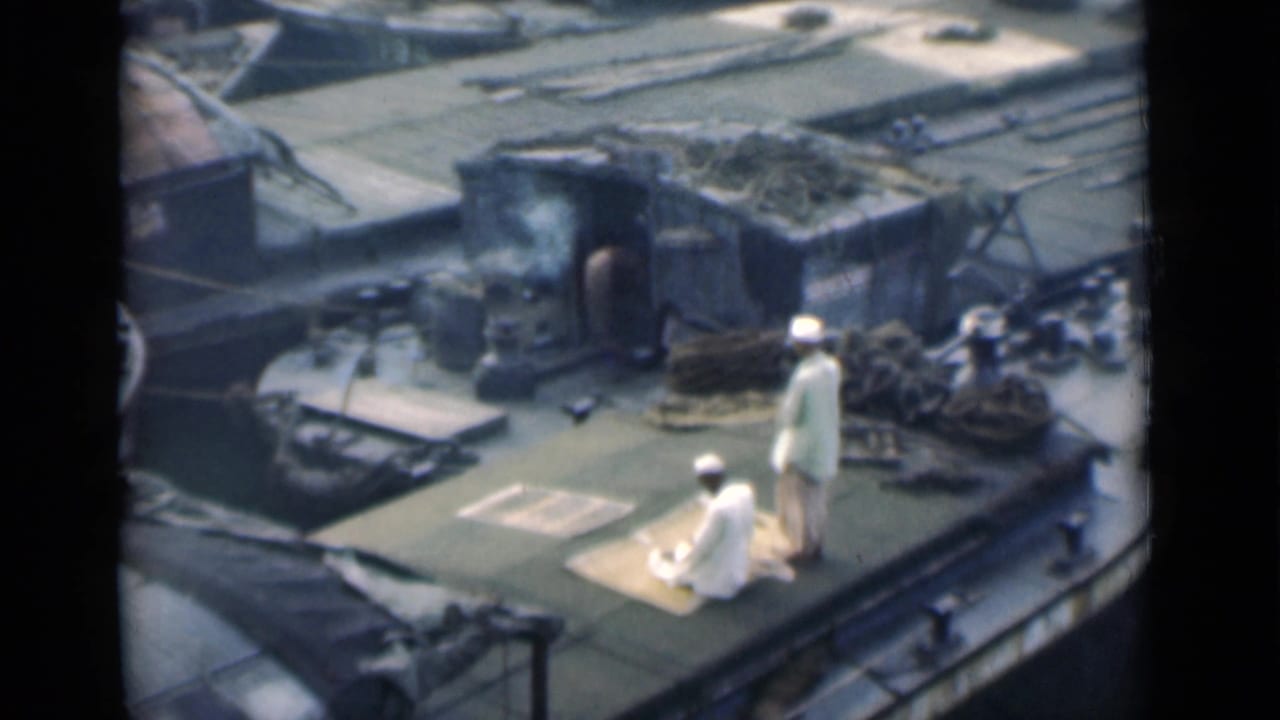 Two men in white clothing on a boat deck, one kneeling on a mat near ropes and cargo.