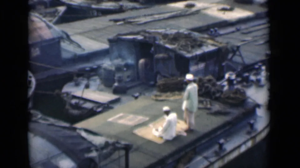 From an elevated home-movie viewpoint, two men in white clothing are seen on a Calcutta boat deck amid cargo, rope, and deck structures.