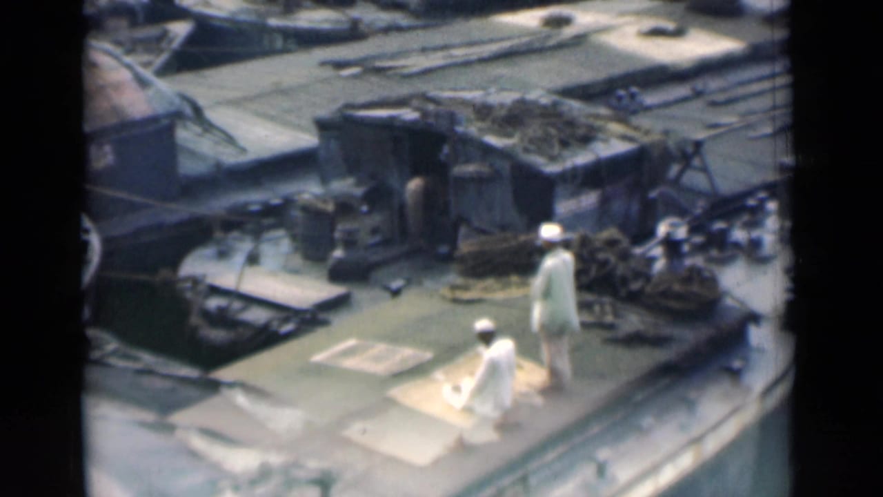 1946 Calcutta 8mm frame showing two men in white traditional clothing on a working boat deck, with one kneeling on a mat near bundled gear.