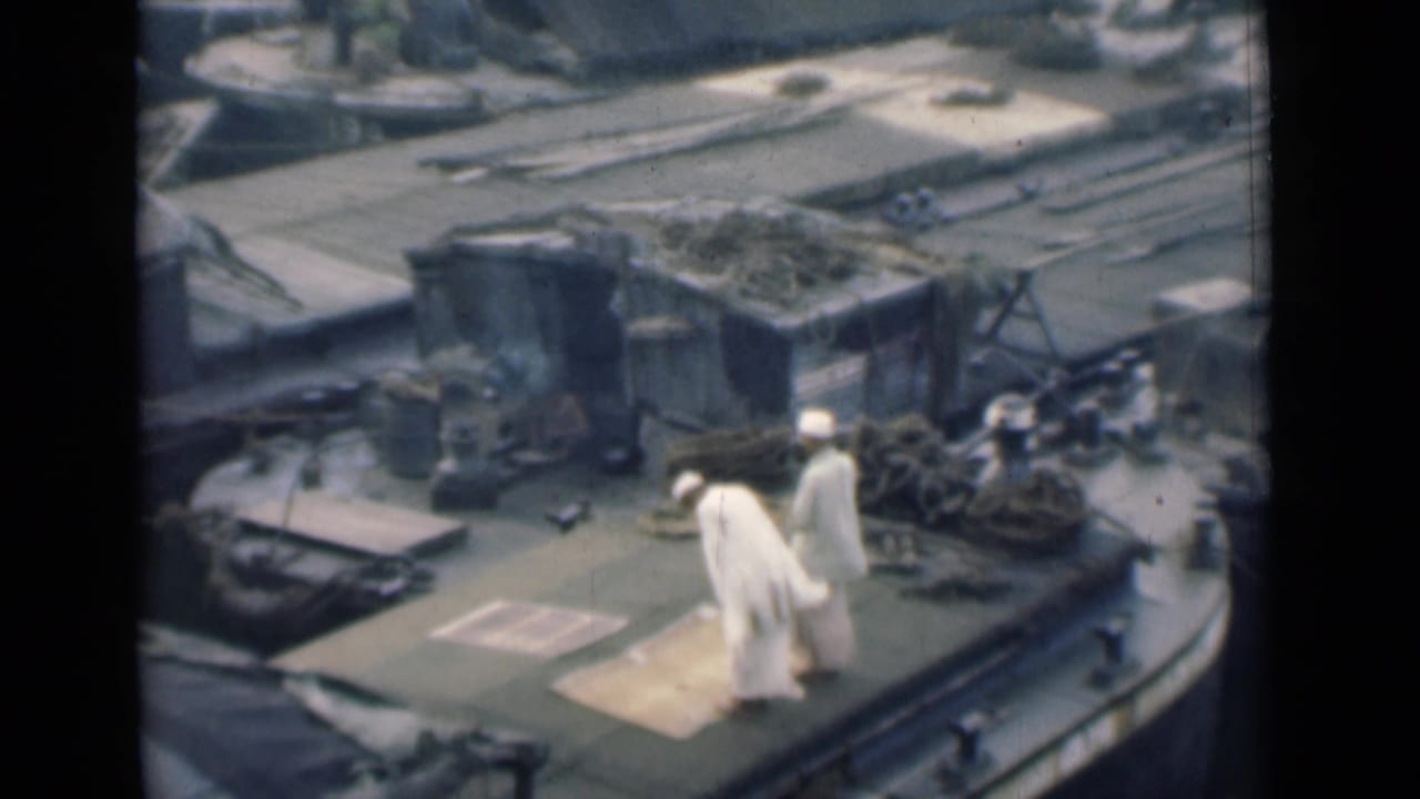 Two men in white garments stand on a boat deck beside a mat and bundled gear.