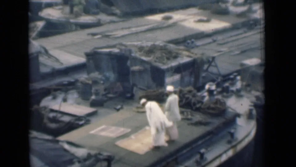 1946 Calcutta 8mm frame with two men in white traditional clothing standing on a vessel deck amid ropes, mats, and port equipment.