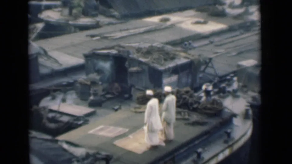 1946 Calcutta home-movie frame showing two men in white clothing crossing a working boat deck with port equipment and river craft nearby.