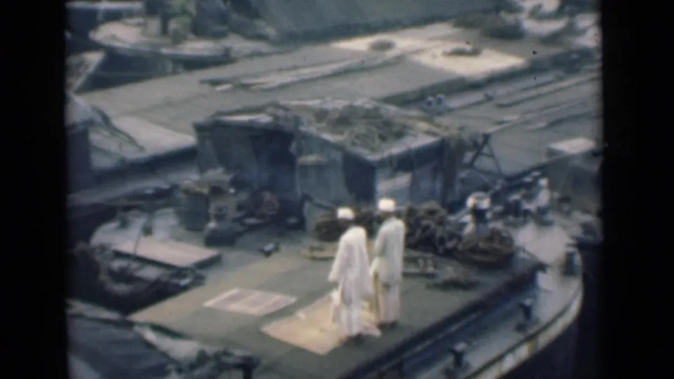 1946 Calcutta 8mm frame showing two men in white traditional clothing near a vessel deck edge, with gear and industrial waterfront shapes behind them.