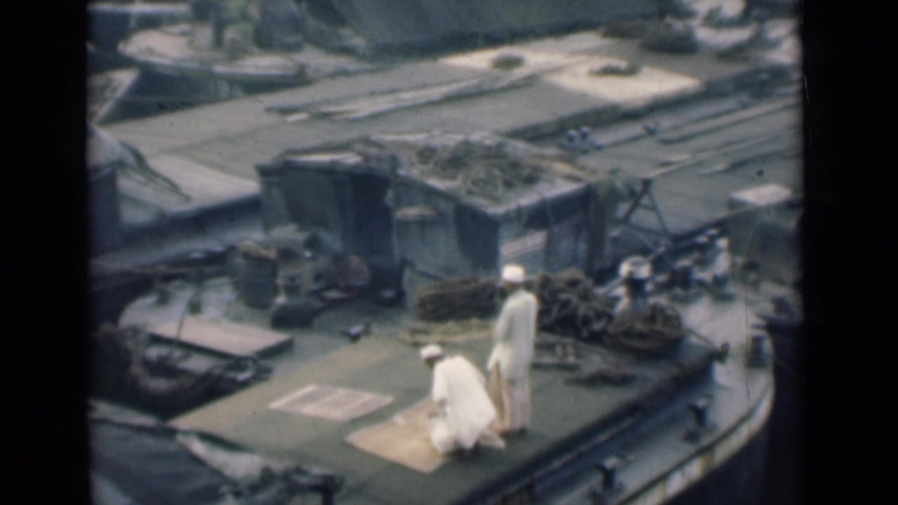 Two men in white clothing on a boat deck near a prayer mat and cargo gear.