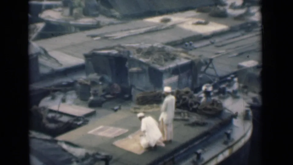 In 1946 Calcutta footage, one man kneels near a mat while another stands on a boat deck with ropes and cargo behind them.