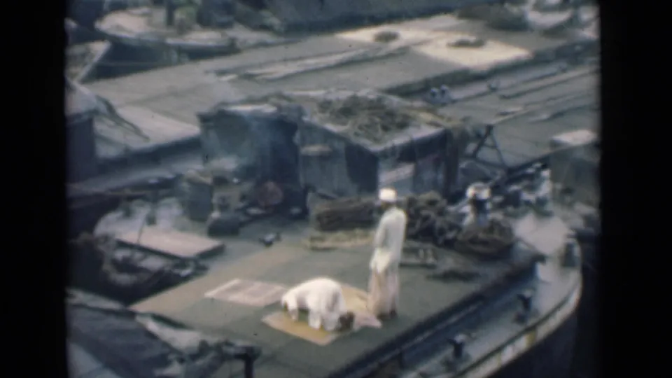 On a working boat deck in Calcutta, a man is prostrate on a prayer mat while another stands near ropes and cargo equipment.