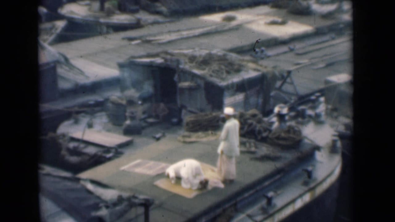 Two men in white clothing pray on a rug laid on a boat deck.
