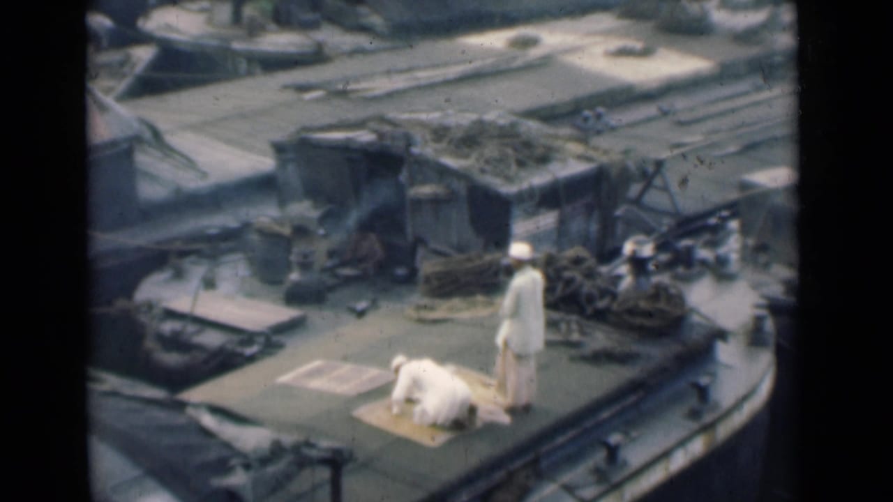 One man kneels on a rug while another stands on a cluttered boat deck.