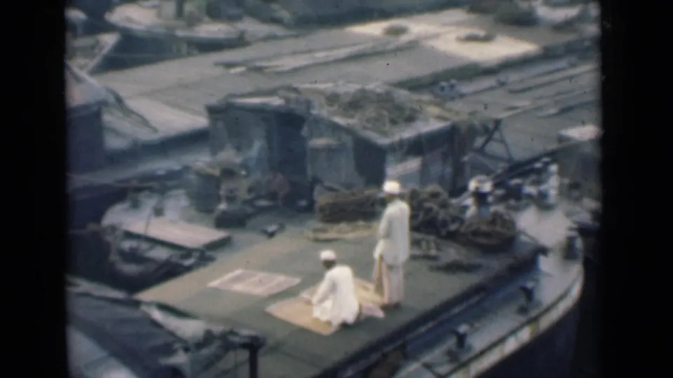 On the boat deck, one man appears to rise or shift position on the rug while the other remains standing in white clothing.
