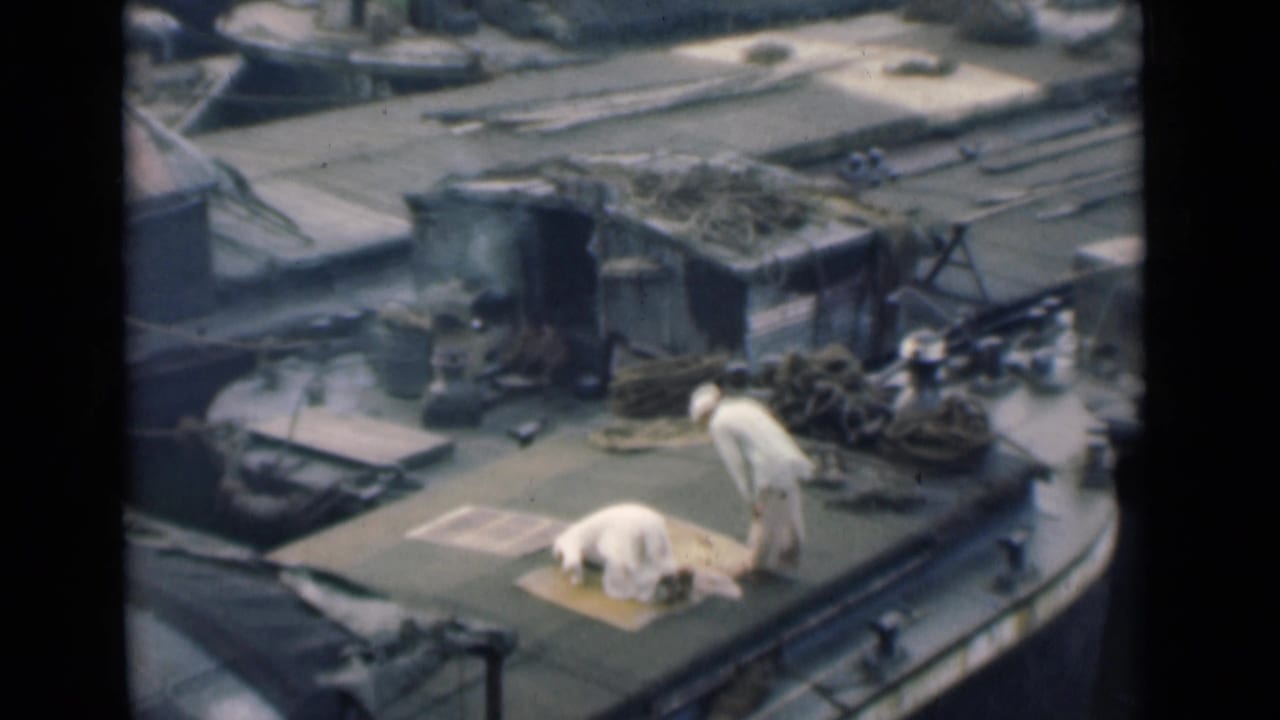Two men in white clothing on a boat deck, one kneeling on a mat and one standing nearby.
